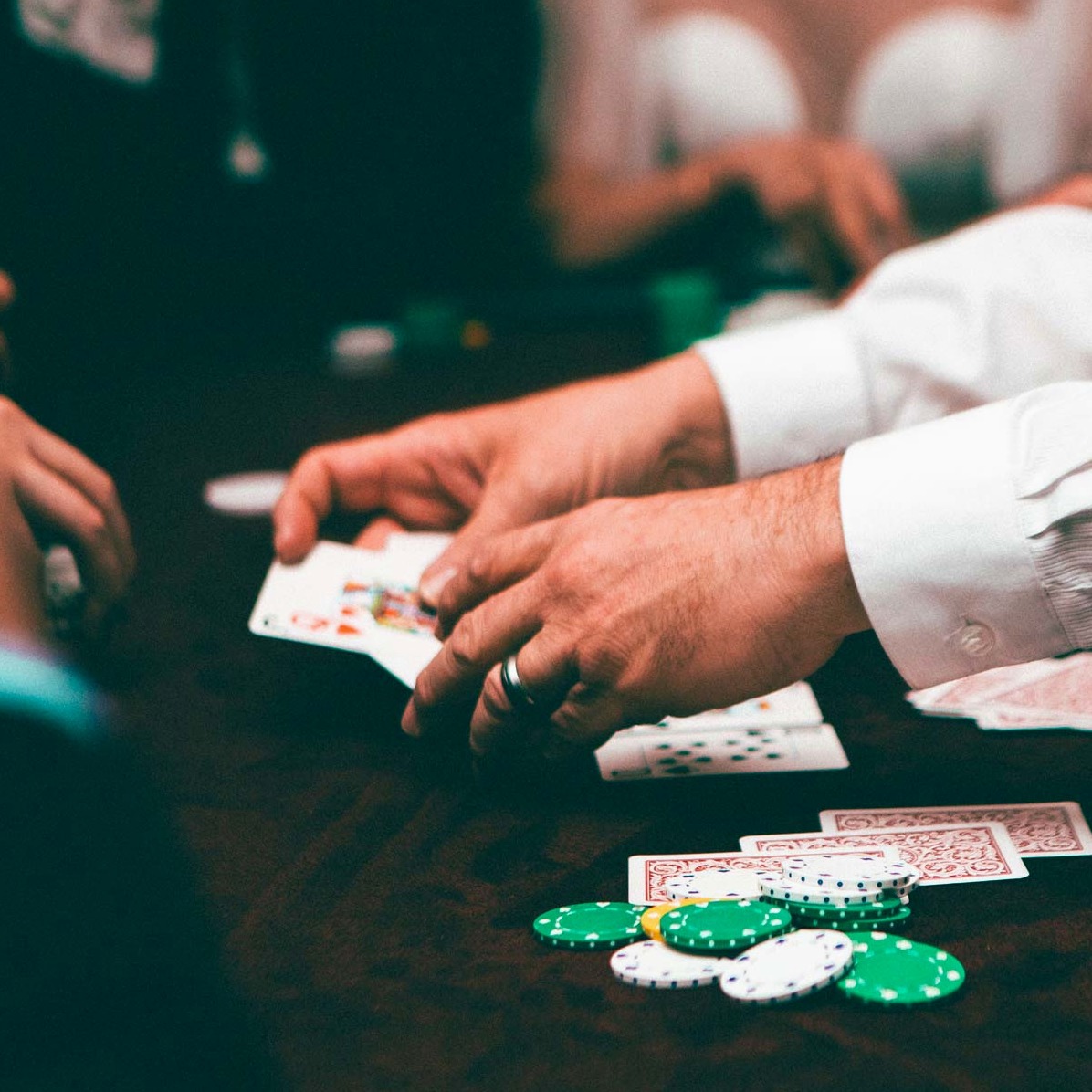 Image of a poker game in progress with four players, including one woman, seated around a wooden table without felt. There are poker chips and cards on the table.